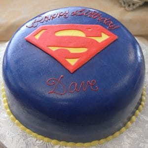 The CB-006E is a round blue cake with a red and yellow Superman logo on top, “Happy Birthday” and “Dave” in red icing, and a yellow bead border around the base.