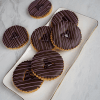 A rectangular white plate holds several CE-000A Chocolate Raspberry Linzer cookies, each round with a chocolate glaze, white drizzle, and a heart-shaped center cutout. Two more cookies rest on the marble surface beside the plate.