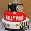 The TC-157 is a three-tier Hollywood-themed birthday cake with gold stars, a film reel, clapperboard featuring "Janai," a silver 16 topper, and "HOLLYWOOD" in white letters on the middle tier.