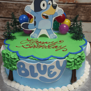 The SC-080 Bluey cake is a round birthday cake with tree and balloon decorations, a big edible Bluey cartoon topper, "Happy Birthday" in red icing, and "BLUEY" in blue letters on the side.