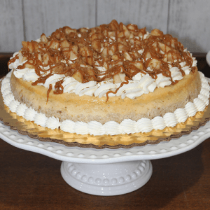 The GD-008 Caramel Apple Cheesecake is topped with whipped cream, caramel drizzle, and nut pieces, and presented on a white decorative cake stand.
