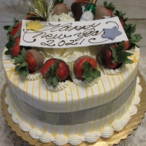 NY-011 Mango Cake: Round cake with white frosting, yellow drizzle, and a silver ribbon. Topped with chocolate-dipped strawberries, a miniature champagne bottle, and a Happy New Year 2021 sign.