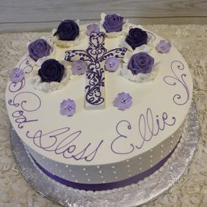 RC-051 is a round cake featuring purple and white roses, a decorative purple cross, and small purple flowers. “God Bless Ellie” is written in purple icing on top, with an elegant white and purple color scheme.