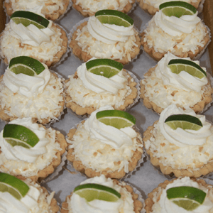 CO-071 Key lime Tarts, each topped with whipped cream, shredded coconut, and a slice of lime, are neatly arranged in rows on a parchment-lined tray.