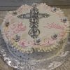 RC-022 is a round white cake with a silver cross, small white flowers, silver dots, ruffled edges, and “God Bless Lillian” in pink writing. It’s presented on an embossed silver board.