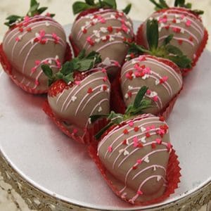 Six VC-033 Milk chocolate dipped strawberries with leafy tops, pink and white drizzle, and heart-shaped sprinkles are arranged on a silver stand with a decorative edge.