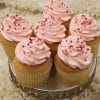 Six VC-038 Display Valentine gold cupcakes, filled with chocolate mousse and topped with light pink frosting and red heart-shaped sprinkles, are arranged on a decorative silver stand against a white textured background.