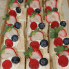 Rows of CO-072 Tres Leches Squares, topped with whipped cream, halved strawberries, blueberries, and raspberries, neatly arranged on a tray.