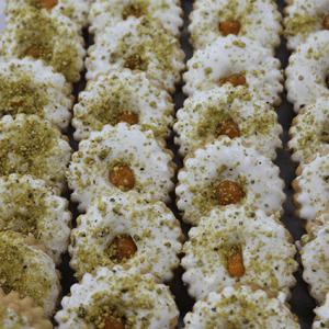 Rows of CO-047 Tea cookies with scalloped edges, dipped in white icing, sprinkled with chopped pistachios, and finished with a dollop of apricot jam in the center.