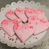 CE-020e Valentine's heart-shaped cookies with pink icing, sprinkles, and "love" written on each are displayed on a white scalloped plate, topped with small heart-shaped confetti.