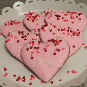 CE-020f Valentine's: Heart-shaped cookies with pink icing and "love," decorated with red, pink, and white heart sprinkles, arranged on a white scalloped plate.