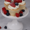 Three CC-000F Tres leche cupcakes with whipped cream, strawberries, and blueberries are on a white cake stand, with extra raspberries, blueberries, and strawberries scattered on a marble surface.
