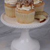 Three CC-000G Apple chai cupcakes with drizzle are displayed on a white pedestal cake stand, accompanied by apple slices on a gray marble surface.
