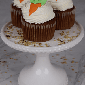 Three CC-000L carrot cupcakes with white frosting and carrot-shaped decorations are displayed on a white cake stand, with crumbs scattered around.