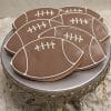 CE-002a sports: Football-shaped sugar cookies with brown and white icing, displayed on a decorative silver cake stand with crystal accents against a textured white background.