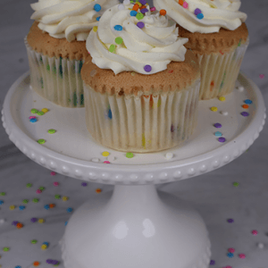 Three CC-000P Funfetti Cupcakes with white frosting and colorful sprinkles sit on a white cake stand, with extra sprinkles scattered around on a gray surface.