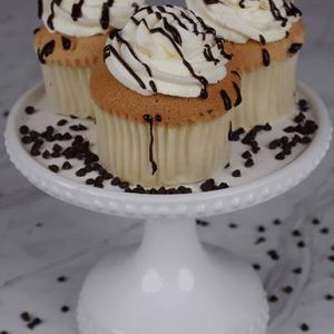 Three CC-007 Display Gold whipped cream Cupcakes with swirled white frosting and chocolate drizzle sit on a white cake stand, surrounded by scattered mini chocolate chips.