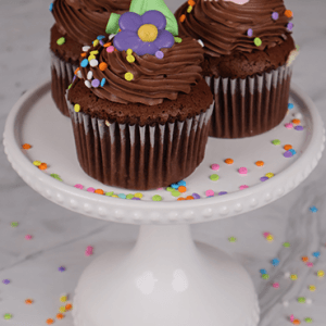 Three CC-001 chocolate cupcakes with white mousse filling, topped with colorful flower decorations and confetti sprinkles, are arranged on a white cake stand with extra sprinkles around.