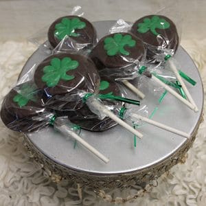 STP-027 Chocolate dipped Oreos, decorated with green shamrock shapes and wrapped in clear plastic, are displayed on a silver stand with a decorative edge on a white textured surface.