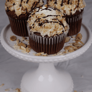 Three CC-000N Almond Joy Cupcakes, topped with whipped cream, toasted coconut, sliced almonds, and chocolate drizzle, are displayed on a white cake stand with almonds scattered around the base.