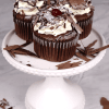 Three CC-000W Black Forest Cupcakes, topped with whipped cream, chocolate shavings, and cherries, are arranged on a white cake stand with extra chocolate shavings sprinkled around.