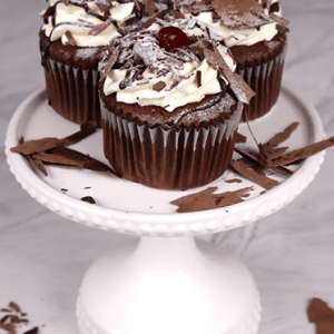 Three CC-000W Black Forest Cupcakes, topped with whipped cream, chocolate shavings, and cherries, are arranged on a white cake stand with extra chocolate shavings sprinkled around.
