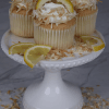 Three CC-000X Lemon Cupcakes topped with whipped cream, toasted coconut flakes, and lemon slices are displayed on a white cake stand, with extra lemon slices and coconut flakes around them.