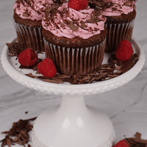 Three CC-000D Chocolate raspberry cupcakes sit on a white cake stand, topped with pink frosting, raspberries, and chocolate decorations. Chocolate shavings and raspberries are scattered around the stand.