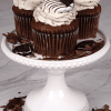 Three CC-000K Chocolate Oreo Cupcakes topped with swirled cookies and cream frosting and half-dipped chocolate sandwich cookies, displayed on a white cake stand with cookie and chocolate pieces scattered around.