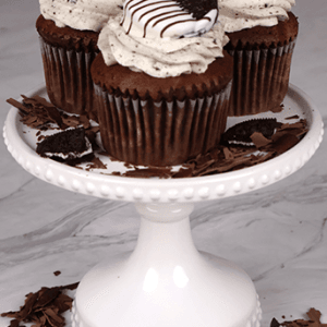 Three CC-000K Chocolate Oreo Cupcakes topped with swirled cookies and cream frosting and half-dipped chocolate sandwich cookies, displayed on a white cake stand with cookie and chocolate pieces scattered around.