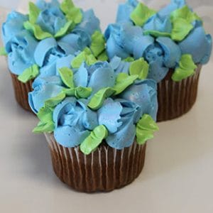 Three CC-090 Chocolate cupcakes with white filling, topped with blue and green petal-shaped icing to resemble hydrangea flowers, are arranged on a white plate for a floral presentation.