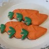 CE-040h Easter: Iced cookies shaped and decorated as carrots, with orange bodies and green tops, arranged on a white scalloped-edge plate.