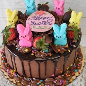 EA-013 is a chocolate frosted cake topped with chocolate-dipped strawberries, marshmallow bunny Peeps, rainbow sprinkles, and a round “Happy Easter” topper surrounded by festive decorations.