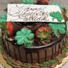 STP-006: Chocolate cake with ganache drip, chocolate-covered strawberries, green shamrock decorations, a white “Happy Birthday Patrick” plaque in brown script, and green sprinkles around the edges.