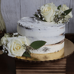 A rustic, semi-naked BR-005 Chocolate cake/chocolate mousse decorated with white roses, greenery, and baby's breath, presented on a gold cake board atop a wooden box against a wooden backdrop.