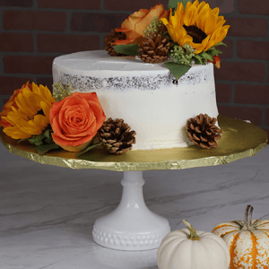 The THX-034 is a white frosted cake on a stand, adorned with sunflowers, an orange rose, green leaves, and pinecones. Beside it on a marble surface are two small pumpkins: one white and one striped.
