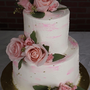 The TC-000K is a two-tiered white cake with pink brushstroke accents, large pink roses, small buds, and green leaves, displayed on a gold cake board before a dark brick backdrop.