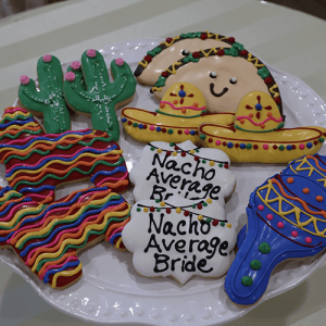 A plate of colorful CE-003i bridal sugar cookies shaped like cacti, tacos, sombreros, piñatas, maracas, and plaques reading “Nacho Average Bride.”.