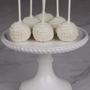 Six DB-000B Gold Cake Pops, each on a stick with gold coating and sprinkles, are arranged on a white beaded-edge cake stand, set on a marble surface against a dark brick background.