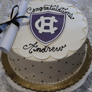 The GC-000S Holy Cross is a round cake with white icing, featuring a purple and white shield emblem, “Congratulations Andrew,” and a diploma tied with a black ribbon, displayed on a white rose-patterned tablecloth.