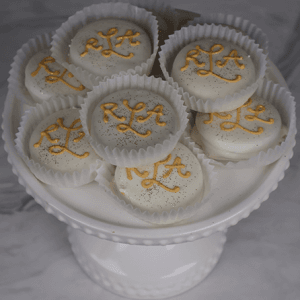 A DB-000H White Dipped Oreo sits in a white cake stand, with eight cookies in paper cups topped with gold sprinkles and yellow iced script, set against a soft gray marble background.