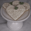 Four DB-000K Bride Cookies, heart-shaped with ornate white icing and small green flower accents, are arranged on a white cake stand against a gray marble background.