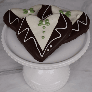 Three DB-000L Groom Cookies shaped like hearts and decorated with white, brown, and green icing to resemble tuxedos are arranged on a white pedestal cake stand against a marble background.
