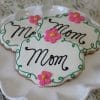 Three CE-050b Mother's Day cookies are on a white plate, each with "Mom" in black icing, green borders, and a pink flower with yellow center. The background is light and textured.