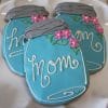 Three blue mason jar-shaped cookies from CE-050a Mother's Day, with "mom" in white icing and pink flower and green leaf decorations, are arranged on a white plate.