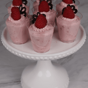 Seven CO-106 Raspberry Shooters—small clear cups of pink mousse topped with a raspberry and chocolate decor—are arranged on a white cake stand against a marble background.