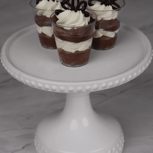 CO-100 Chocolate Shooters: Three small cups of layered chocolate and cream mousse, topped with whipped cream and chocolate pieces, arranged on a white beaded cake stand against a marble background.