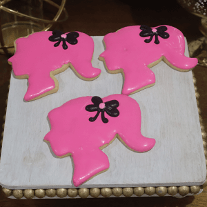 Three "CE-001f birthday" cookies shaped like a girl's head with a ponytail, pink icing, and black bows are displayed on a white wooden tray.