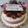 The JC-009 Gold/Mango is a festive cake topped with chocolate-dipped strawberries, American flags, and a “Happy July 4th” card. It features whipped cream, chocolate shavings, and a red, white, and blue ribbon around the base.