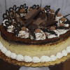 The GD-015 Tiramisu Cheesecake is a round layered cake topped with whipped cream, chocolate shavings, cocoa-dusted candies, and chocolate decorations, presented on a gold cake board against a wooden background.